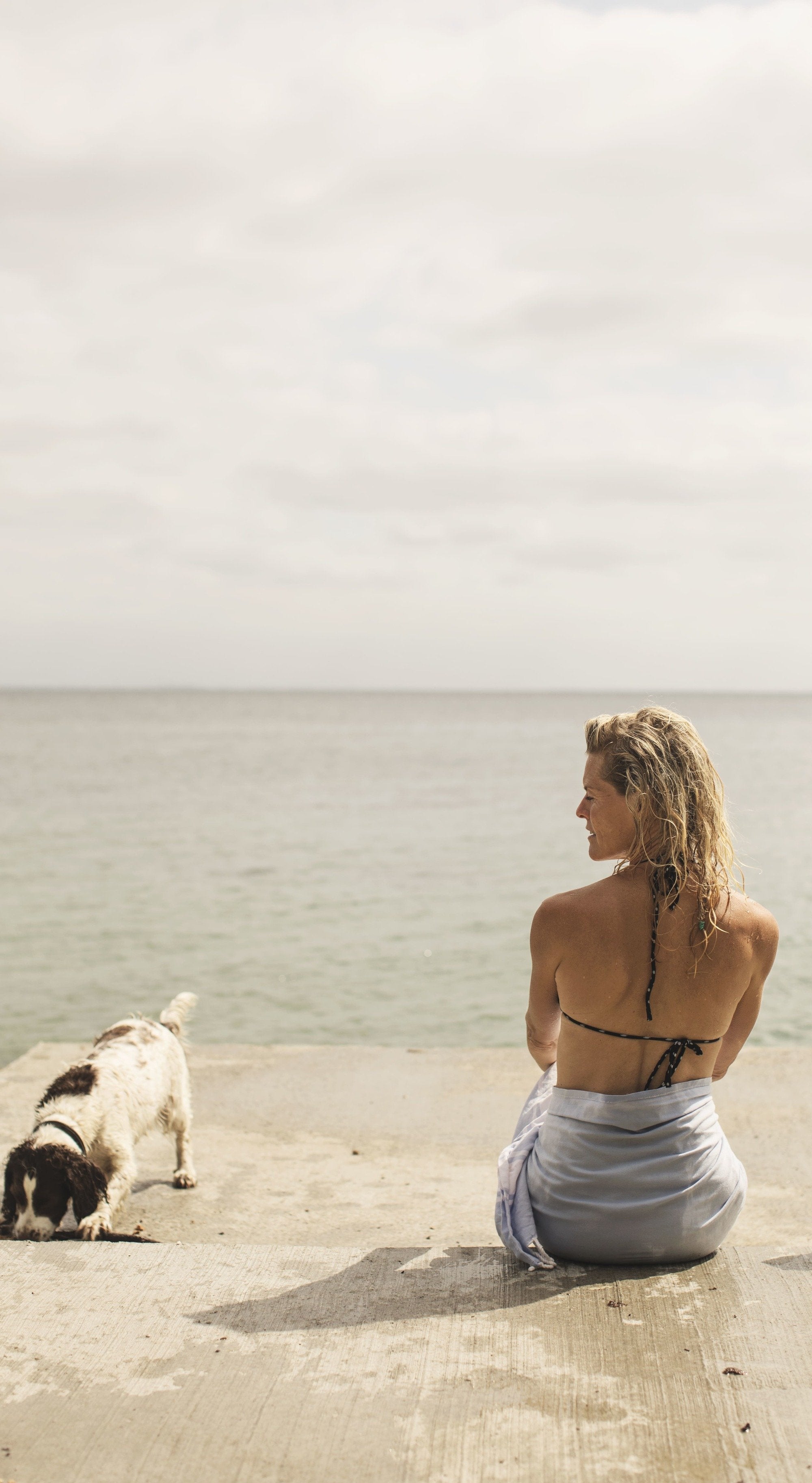 Woman sitting on a stone pier looking out at a calm sea with a small dog nearby, overcast sky.